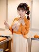 A woman in an orange shirt and white skirt posing in a kitchen.