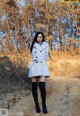 A woman in a white coat and black boots standing on a dirt road.