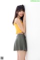 A woman in a yellow top and green skirt leaning against a wall.